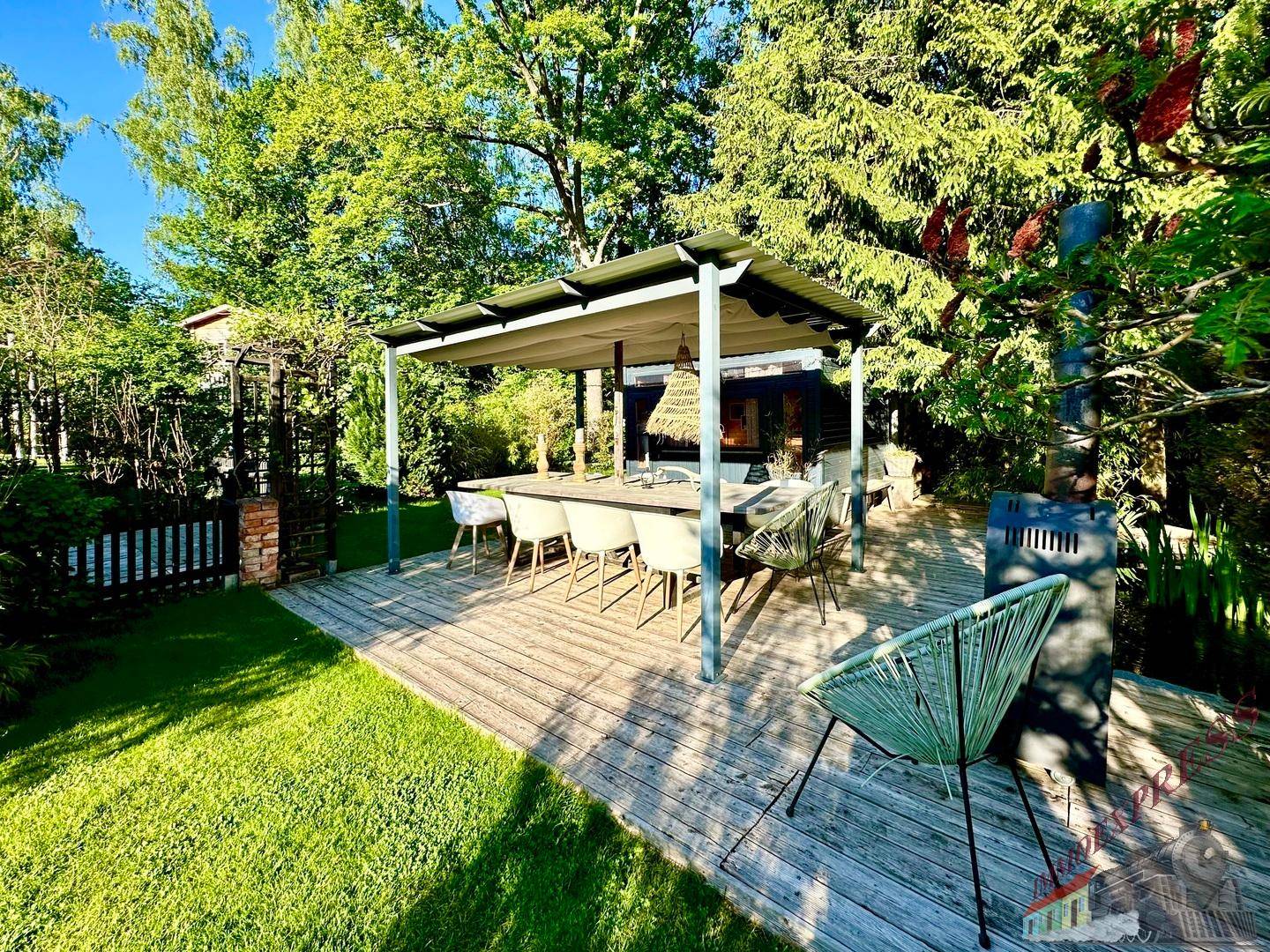 Überdachte Holzterrasse mit großem Esstisch und Stühlen, umgeben von Bäumen.