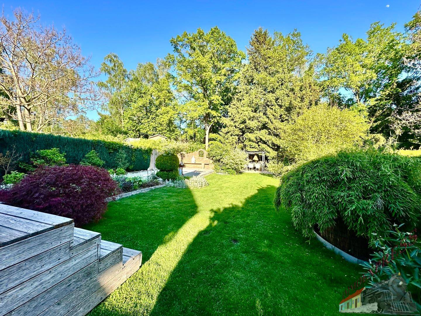 Weitläufiger Garten mit Rasen, Bäumen und Holzterrasse unter strahlendem Himmel.