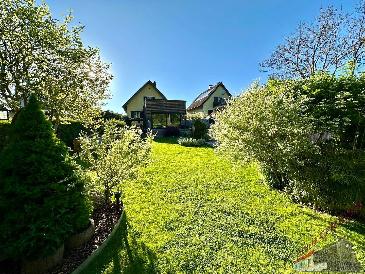Gepflegter Garten mit Rasenfläche, Bäumen und Blick auf das Haus unter blauem Himmel.