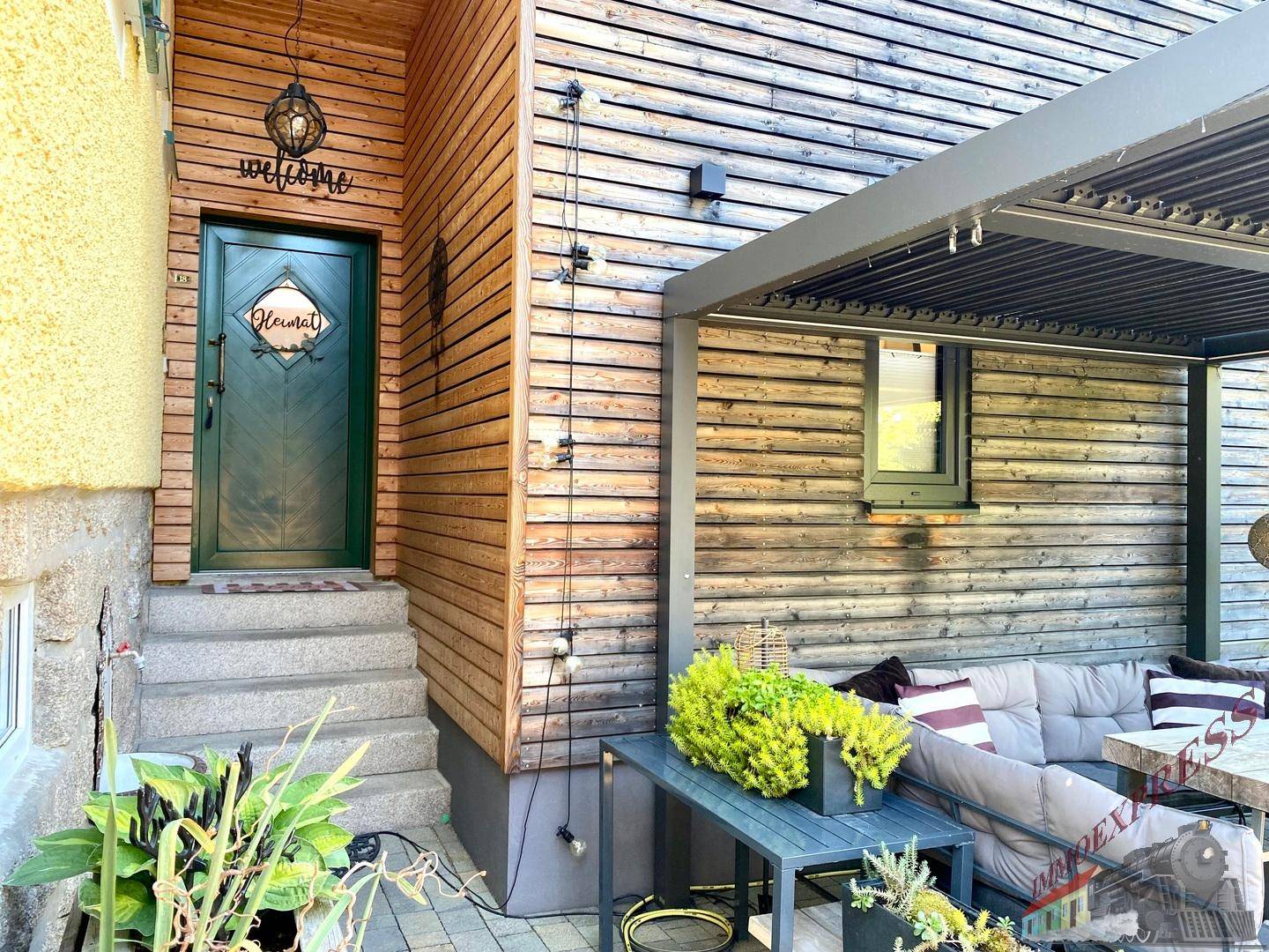 Eingangsbereich mit Holzfassade und überdachter Terrasse mit Sitzgelegenheiten.