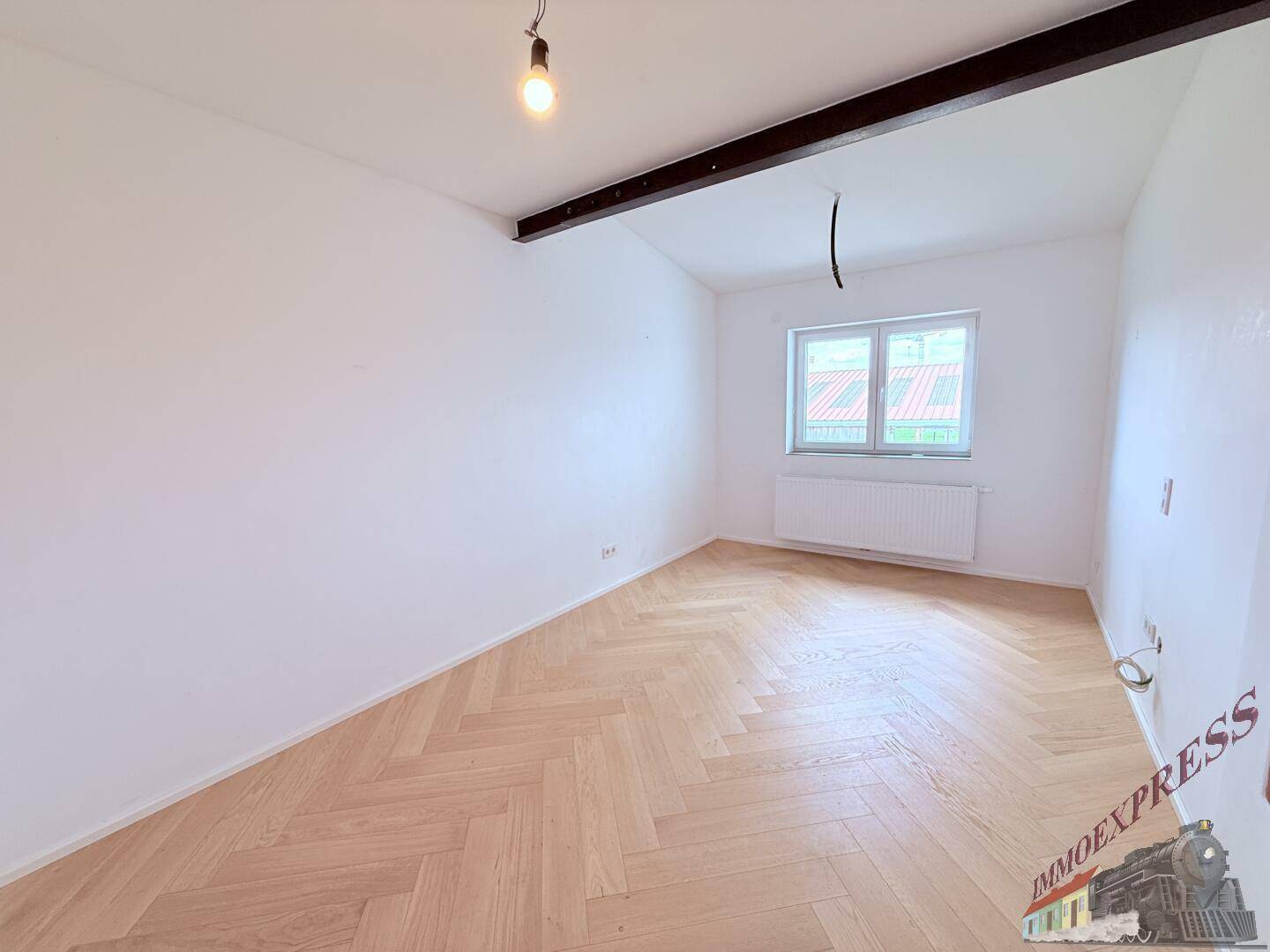 Heller, leerer Raum mit Fischgrätparkett, weißen Wänden und einem Fenster mit Blick nach draußen.