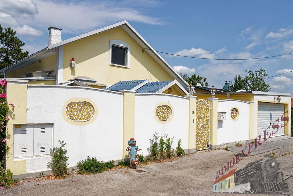 Gelbe Hausfassade mit dekorativem Zaun, Tor und Garageneinfahrt unter blauem Himmel.