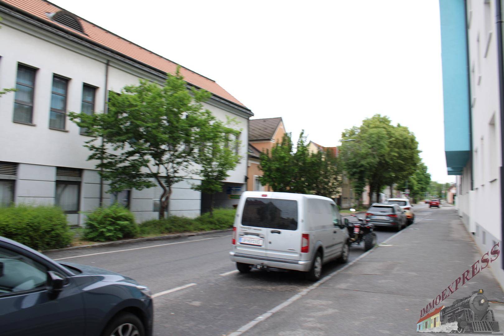 Blick auf eine städtische Straße mit Gebäuden, parkenden Autos und Bäumen entlang des Gehwegs.