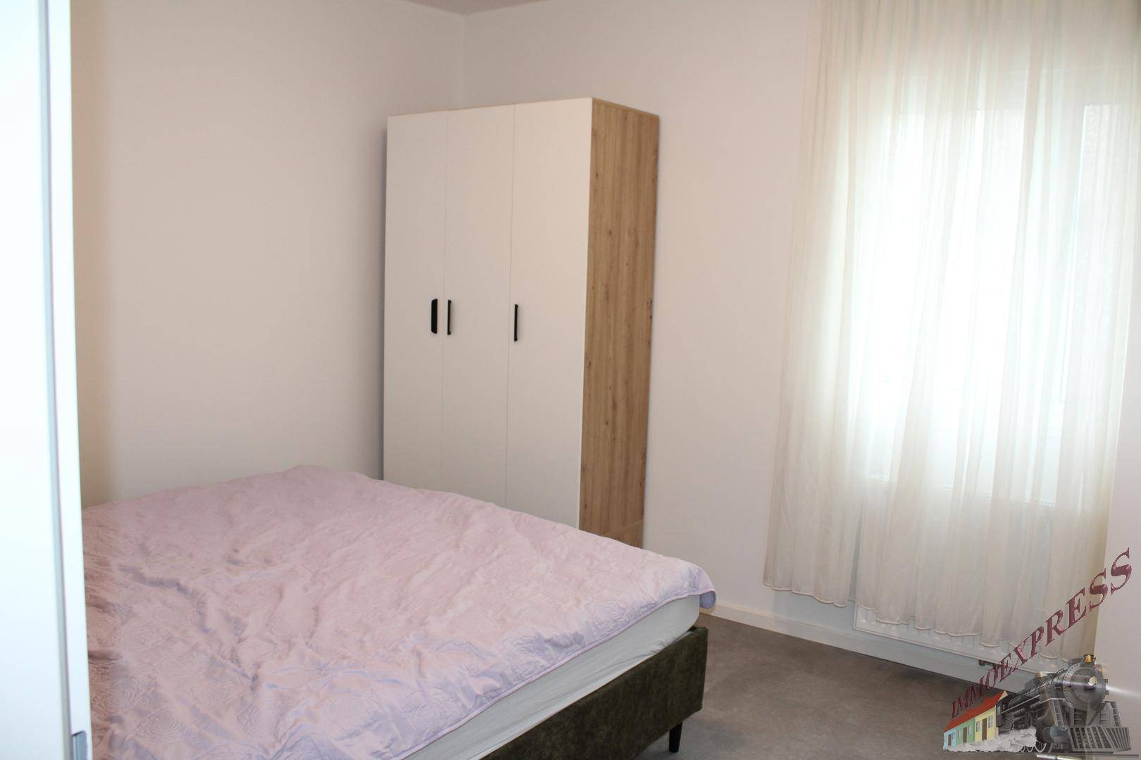 Einfaches Schlafzimmer mit Bett, Kleiderschrank und großem Fenster für viel Tageslicht.