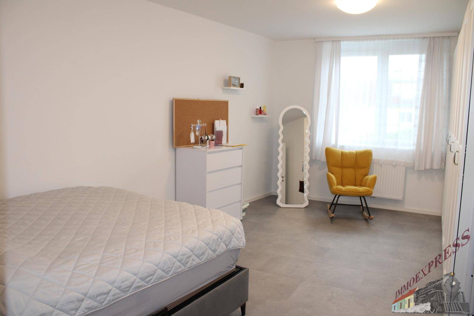 Helles Schlafzimmer mit Doppelbett, Kommode und einem gelben Sessel am Fenster.