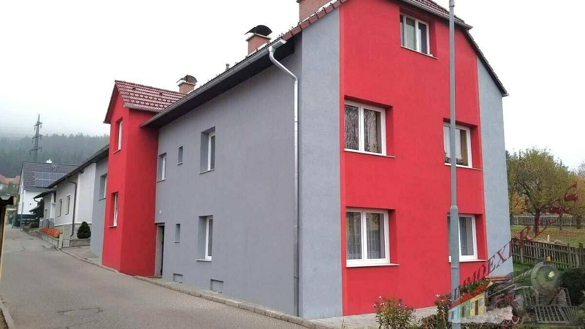 Seitenansicht des Gebäudes mit rot-grauer Fassade, Fenstern und Zufahrt in einer ruhigen Umgebung.