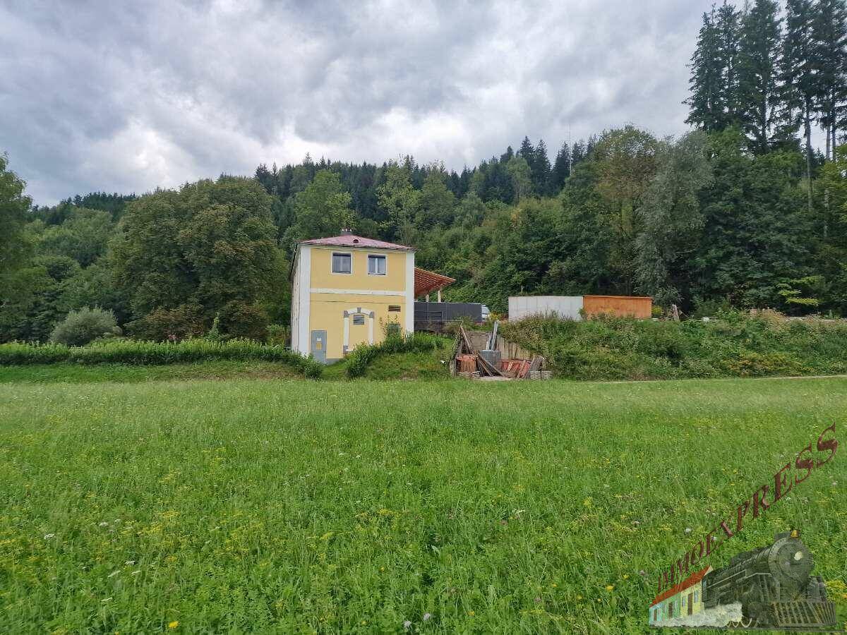 Ein gelbes Gebäude steht inmitten einer weiten grünen Wiese, umgeben von dicht bewaldeten Hügeln.
