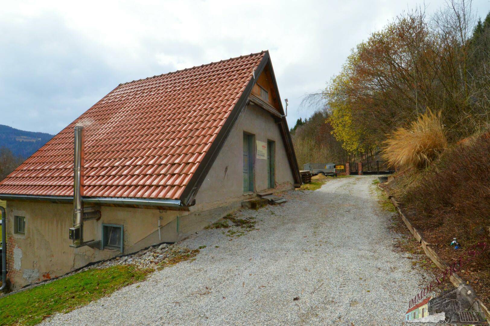 Rustikales Gebäude mit rotem Ziegeldach und Metallkamin, umgeben von einer Schotterauffahrt.