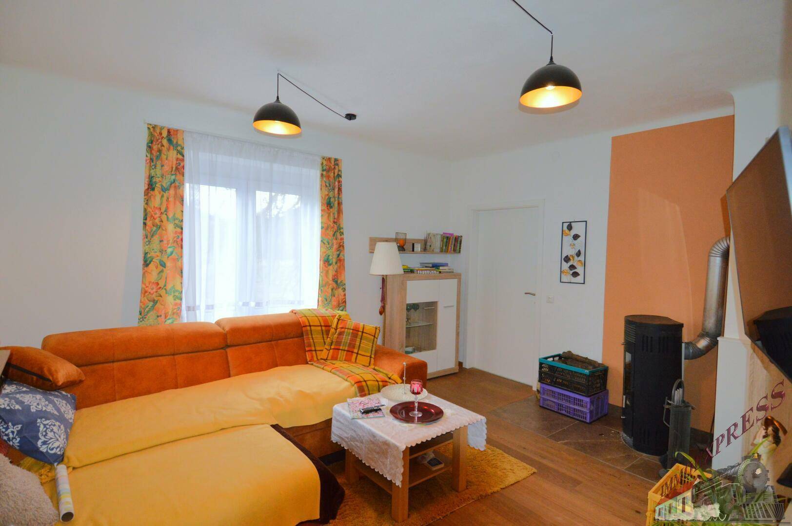 Helles Wohnzimmer mit großem Sofa, Holzofen und Fenster mit Vorhängen.