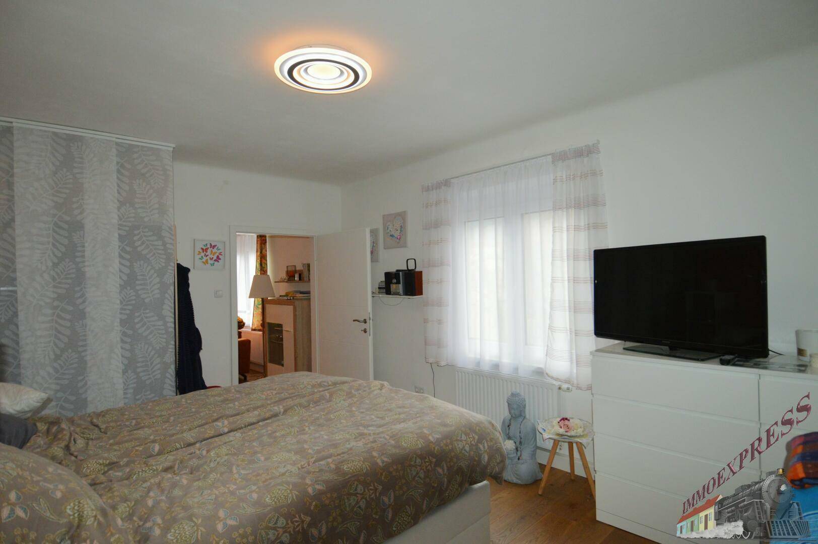 Helles Schlafzimmer mit Doppelbett, Fernseher und Zugang zu einem angrenzenden Raum.