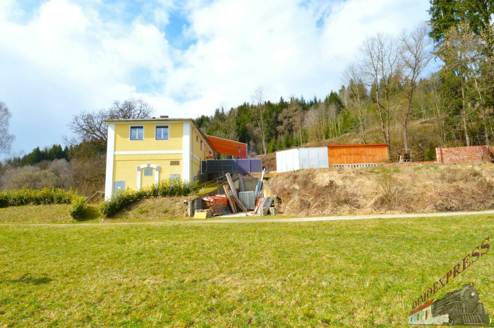 Gelbes Gebäude in ländlicher Umgebung mit Hügeln und Bäumen im Hintergrund.