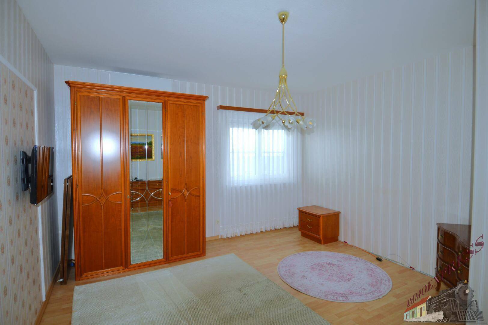 Schlafzimmer mit hellem Holzboden, gemusterten Tapeten und einem großen Holzschrank.