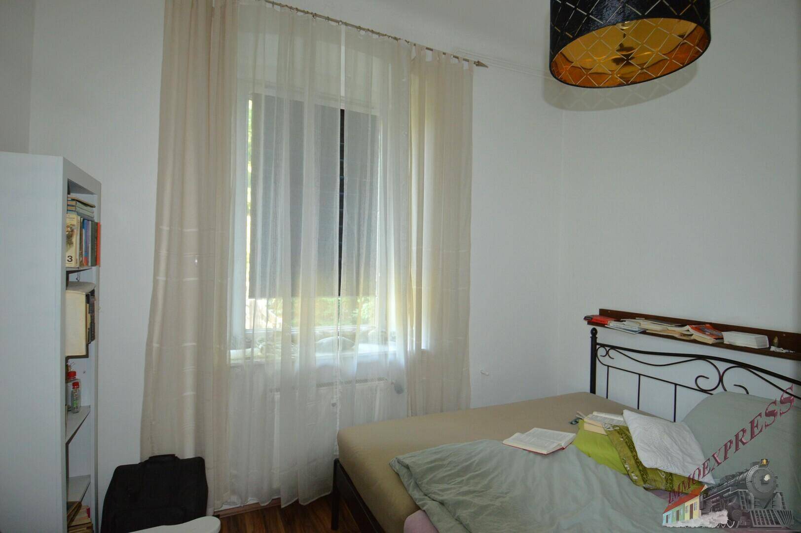 Schlafzimmer mit einem Bett, einem Bücherregal und einem großen Fenster mit Vorhängen.
