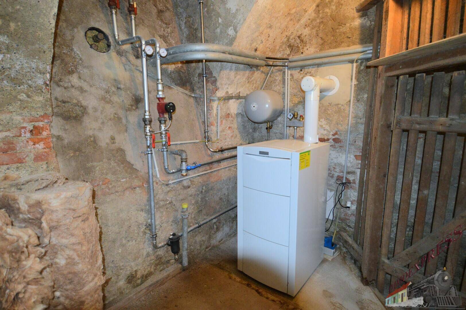 Heizungsraum im Keller mit einem modernen Heizkessel, zahlreichen Rohren und unverputzten Wänden.