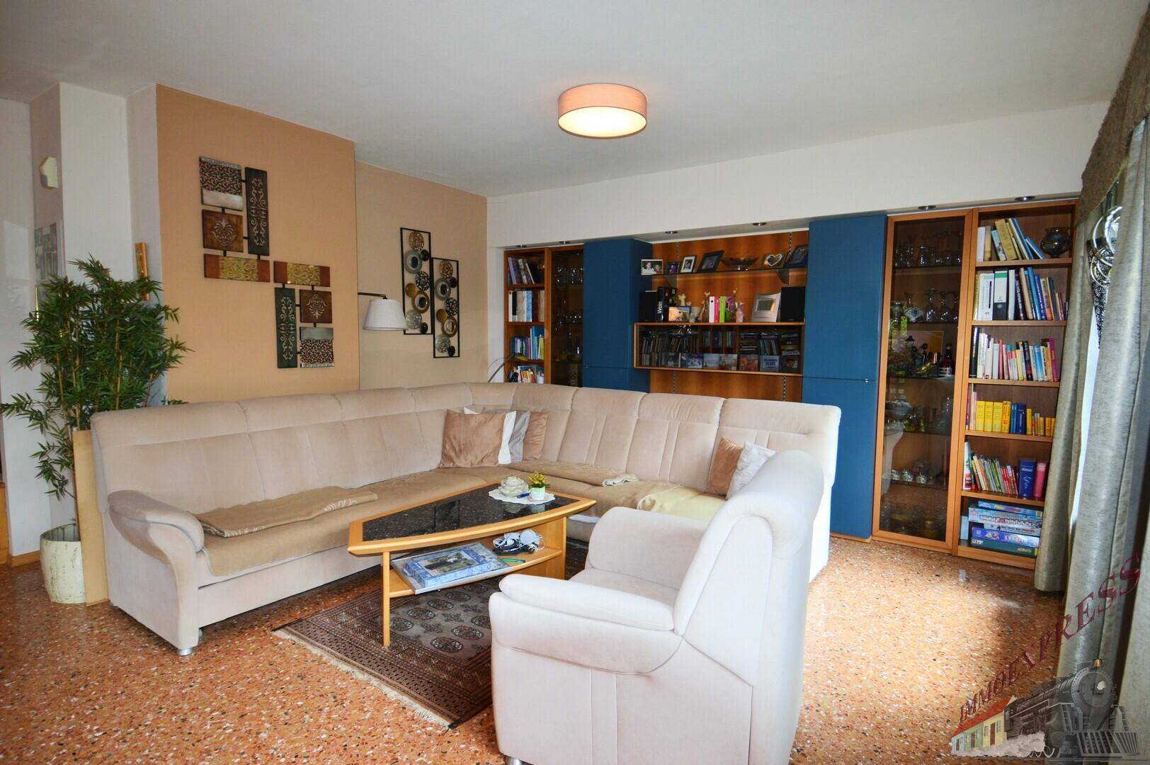 Gemütliches Wohnzimmer mit großem Ecksofa, Terrazzoboden und integrierten Bücherregalen.