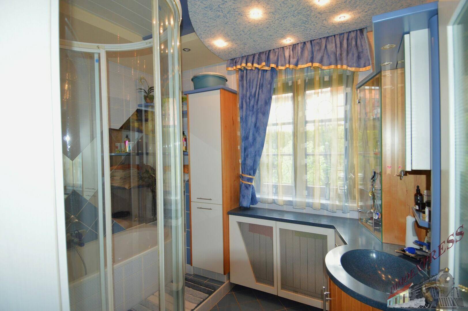 Helles Badezimmer mit Duschkabine, Waschtisch, Fenster und blauen Vorhängen.