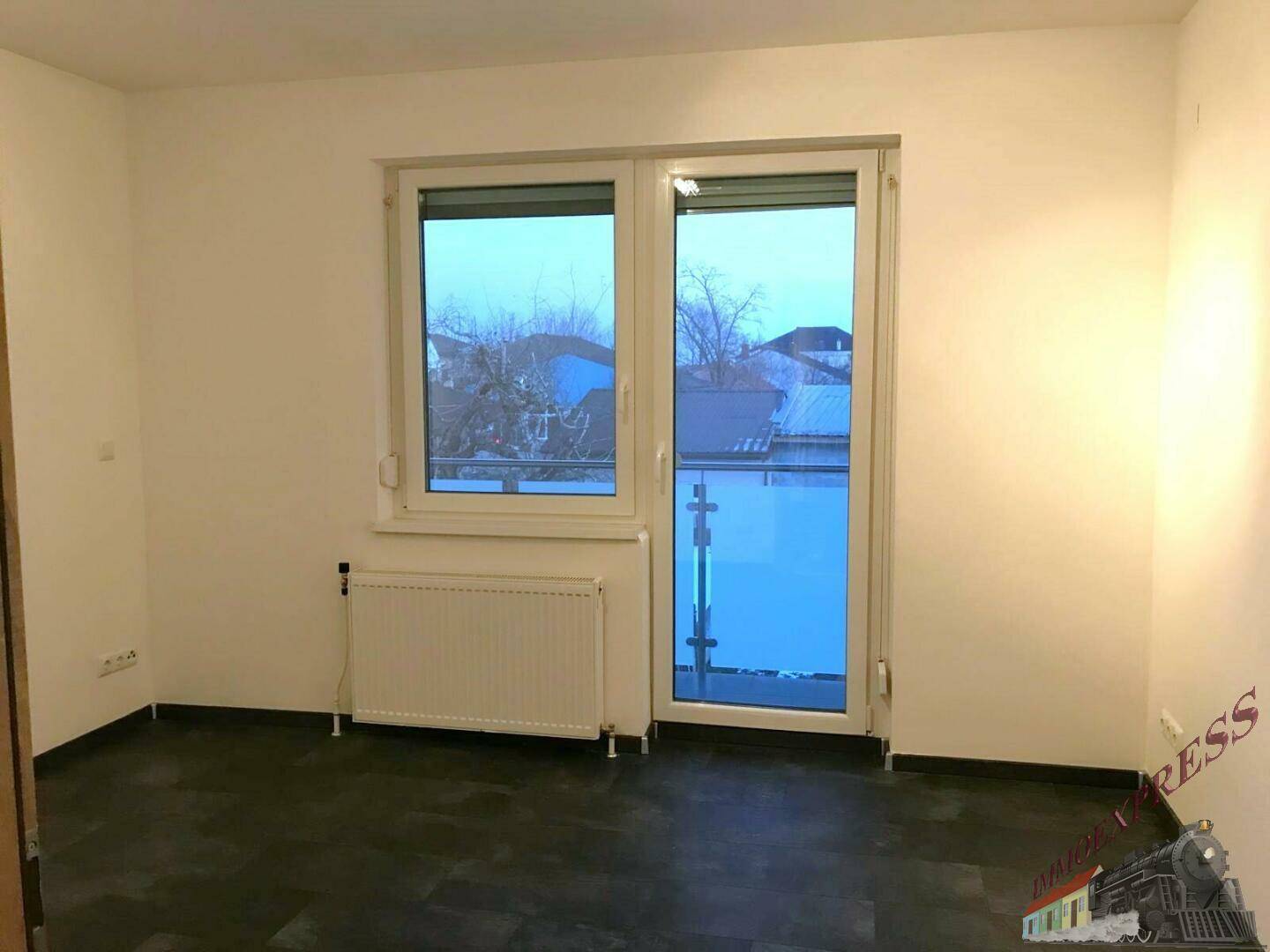 Leerer Raum mit Fenster und Balkonzugang, ausgestattet mit Heizkörper und dunklem Boden.