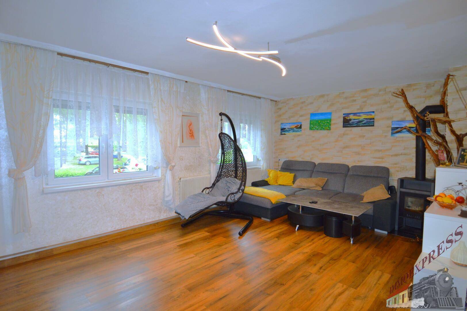 Gemütliches Wohnzimmer mit Holzboden, Kaminofen und einem Hängesessel am Fenster.
