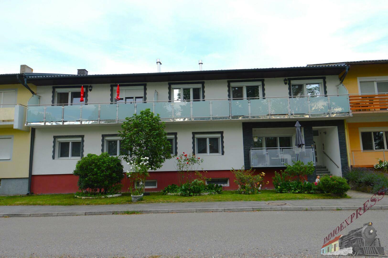Mehrfamilienhaus mit mehreren Balkonen und einer gepflegten Vorgartenfläche an einer Straße.