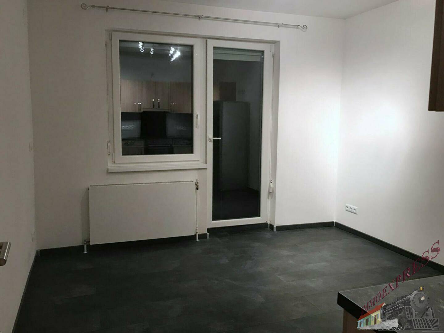 Unmöblierter Raum mit dunklem Boden, Heizkörper und Blick in eine angrenzende Küche.