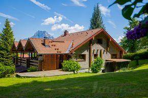 Haus mit großem Rasen und Bergblick, umgeben von grünen Bäumen unter strahlend blauem Himmel.