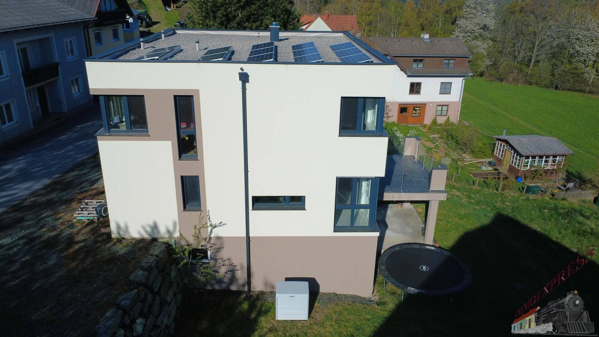 Moderne Hausfassade mit Flachdach, Solaranlage und Trampolin im Gartenbereich.