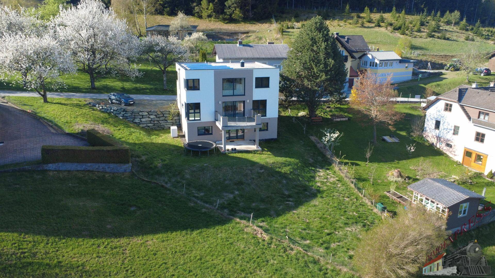 Luftaufnahme eines modernen Hauses mit Flachdach und Solaranlage, eingebettet in eine grüne Hügellandschaft.