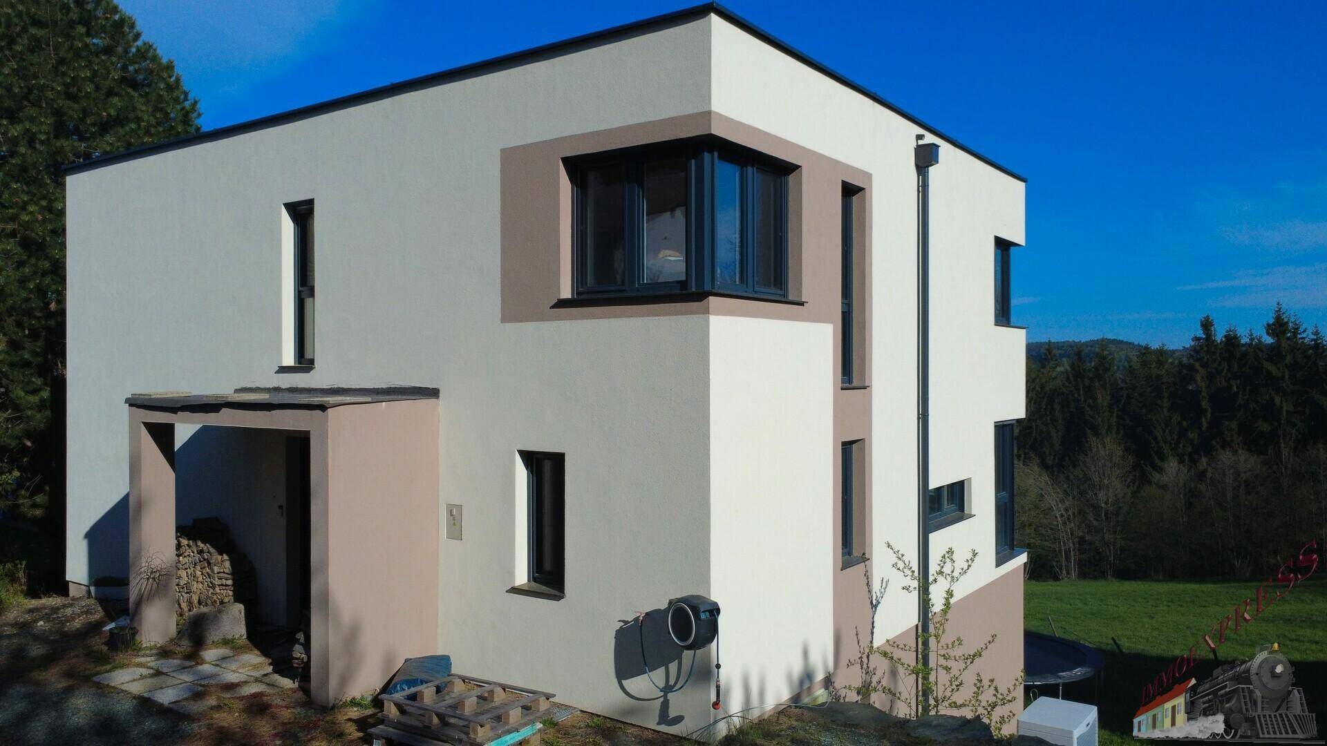 Seitenansicht eines modernen Hauses mit weißer und beiger Fassade, umgeben von Bäumen und Natur.
