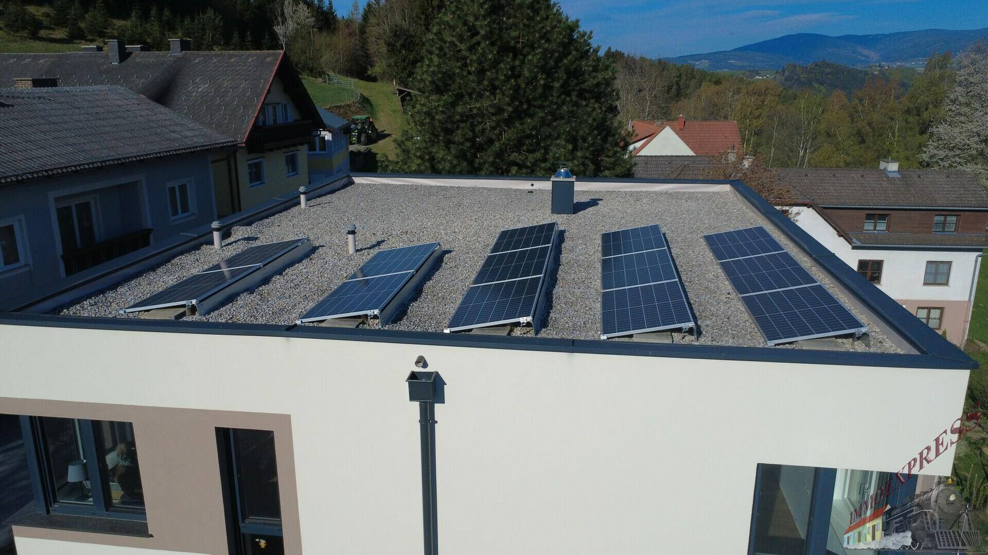 Draufsicht auf das Flachdach mit Photovoltaikanlage und die ländliche Umgebung.