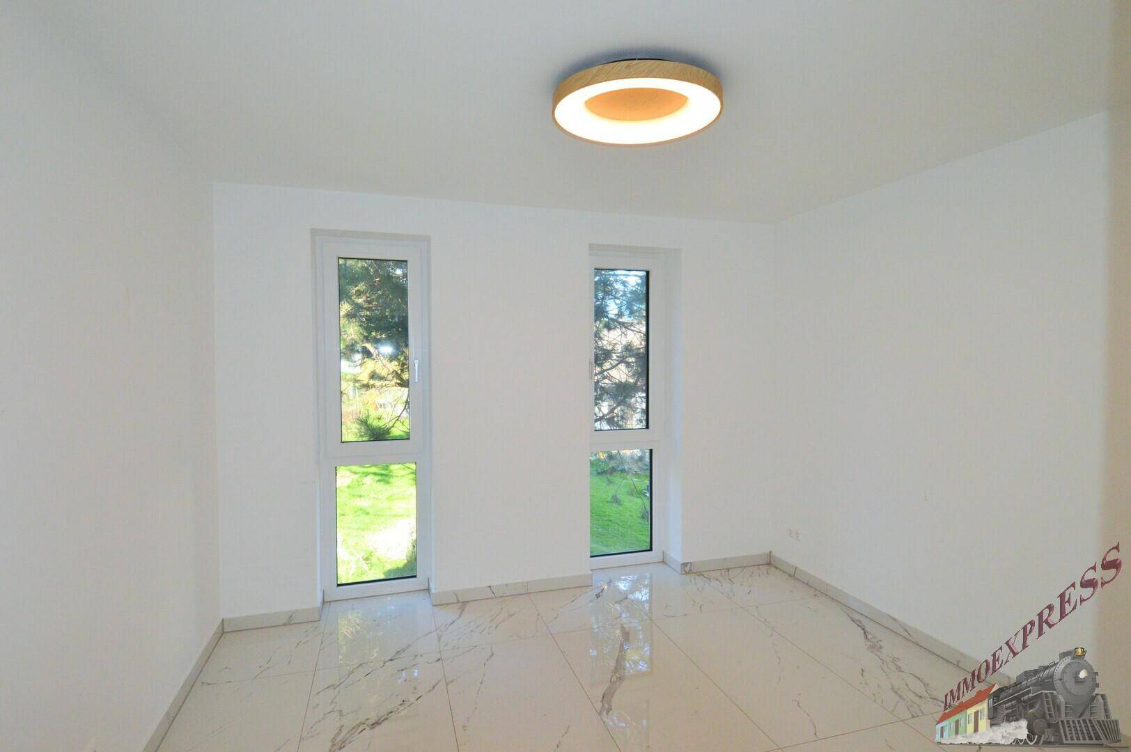 Helles Schlafzimmer mit weißen Wänden, modernen Fliesen und Blick ins Grüne durch die Fenster.