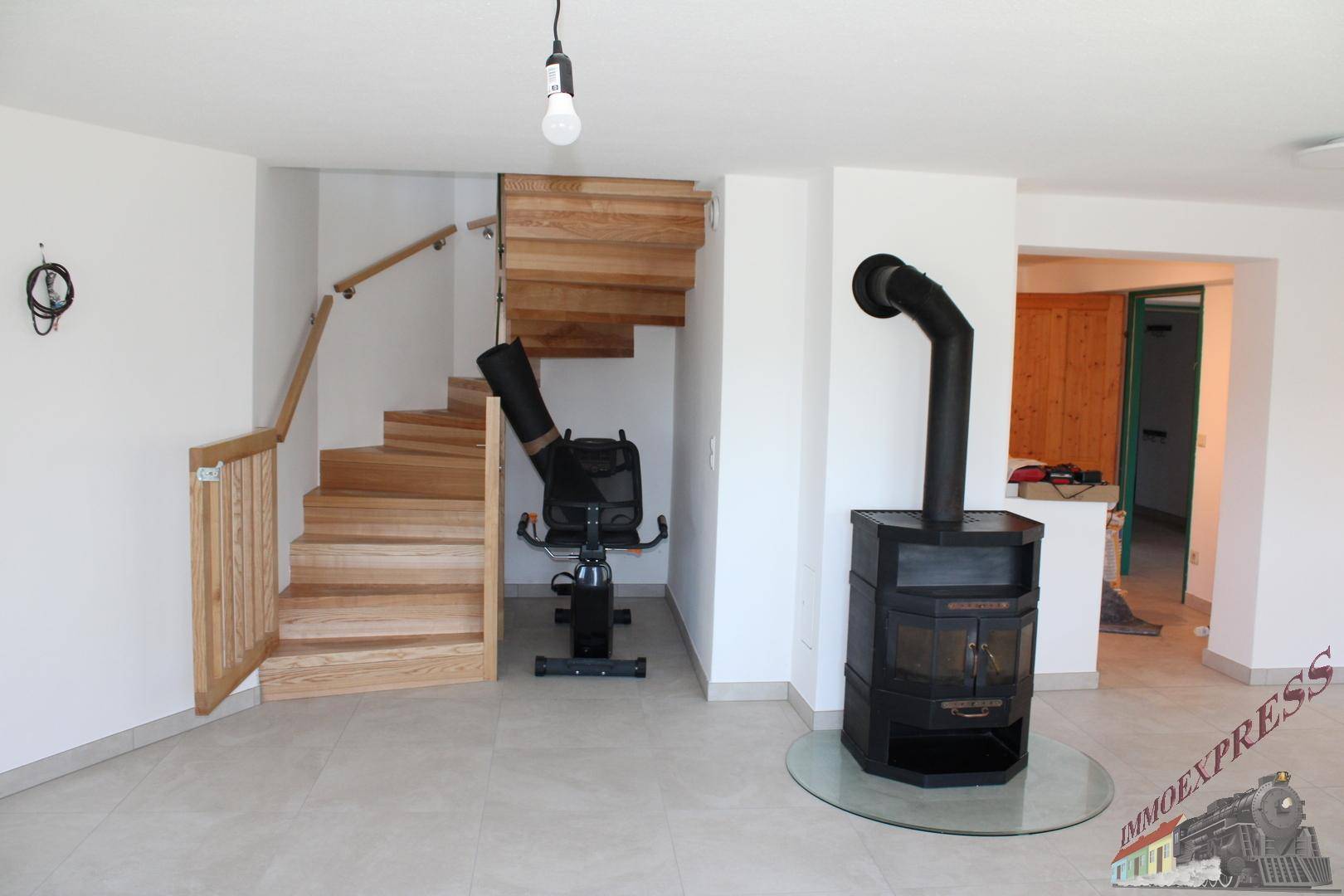 Heller Wohnbereich mit modernem Holzofen, Fliesenboden und einer eleganten Holztreppe.