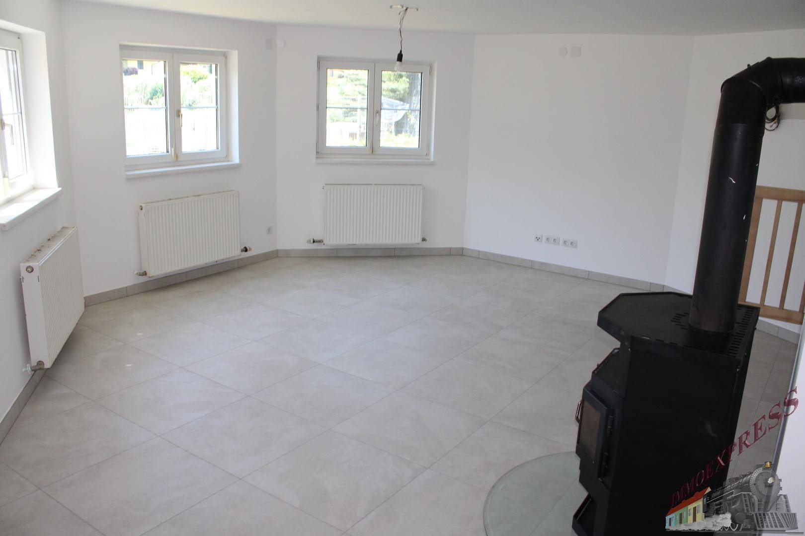 Geräumiges, leeres Wohnzimmer mit Fliesenboden, weißen Wänden und einem Holzofen.