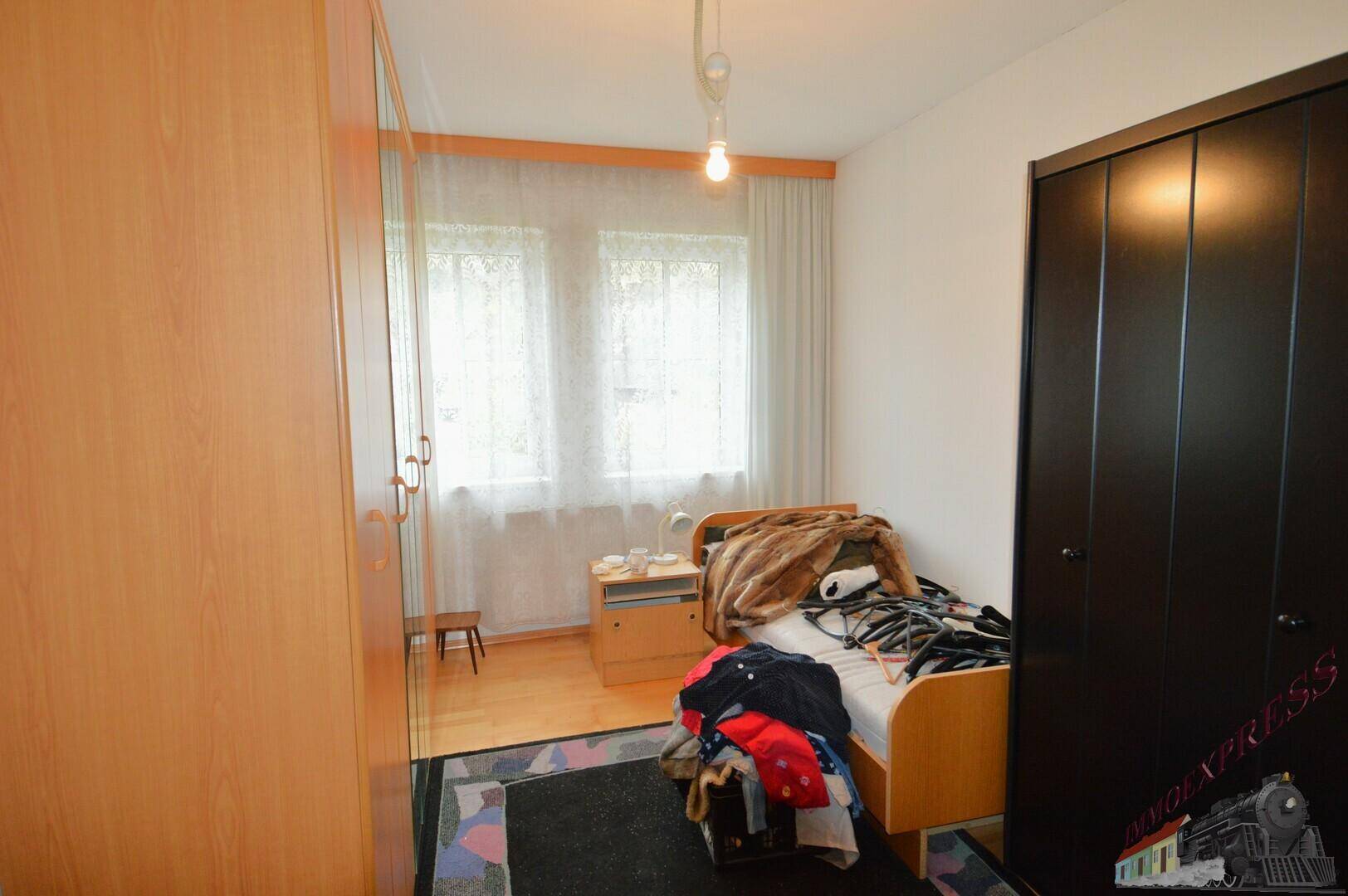 Schlafzimmer mit hellem Holzboden, zwei Fenstern und einfacher Einrichtung, inklusive Kleiderschränken.