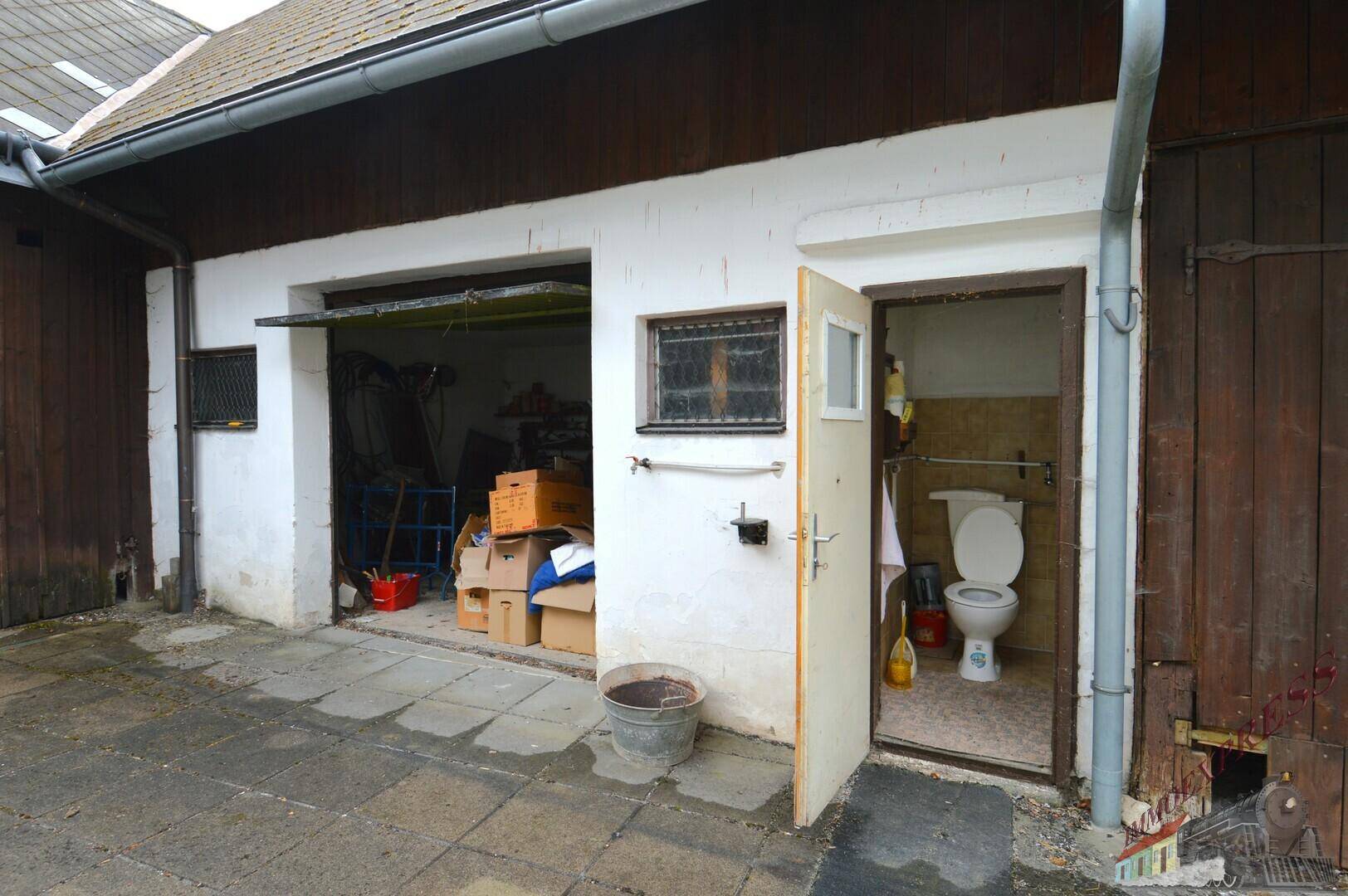 Außenansicht mit Garagentor und einer kleinen Toilette im Freien, daneben ein Holzschuppen.