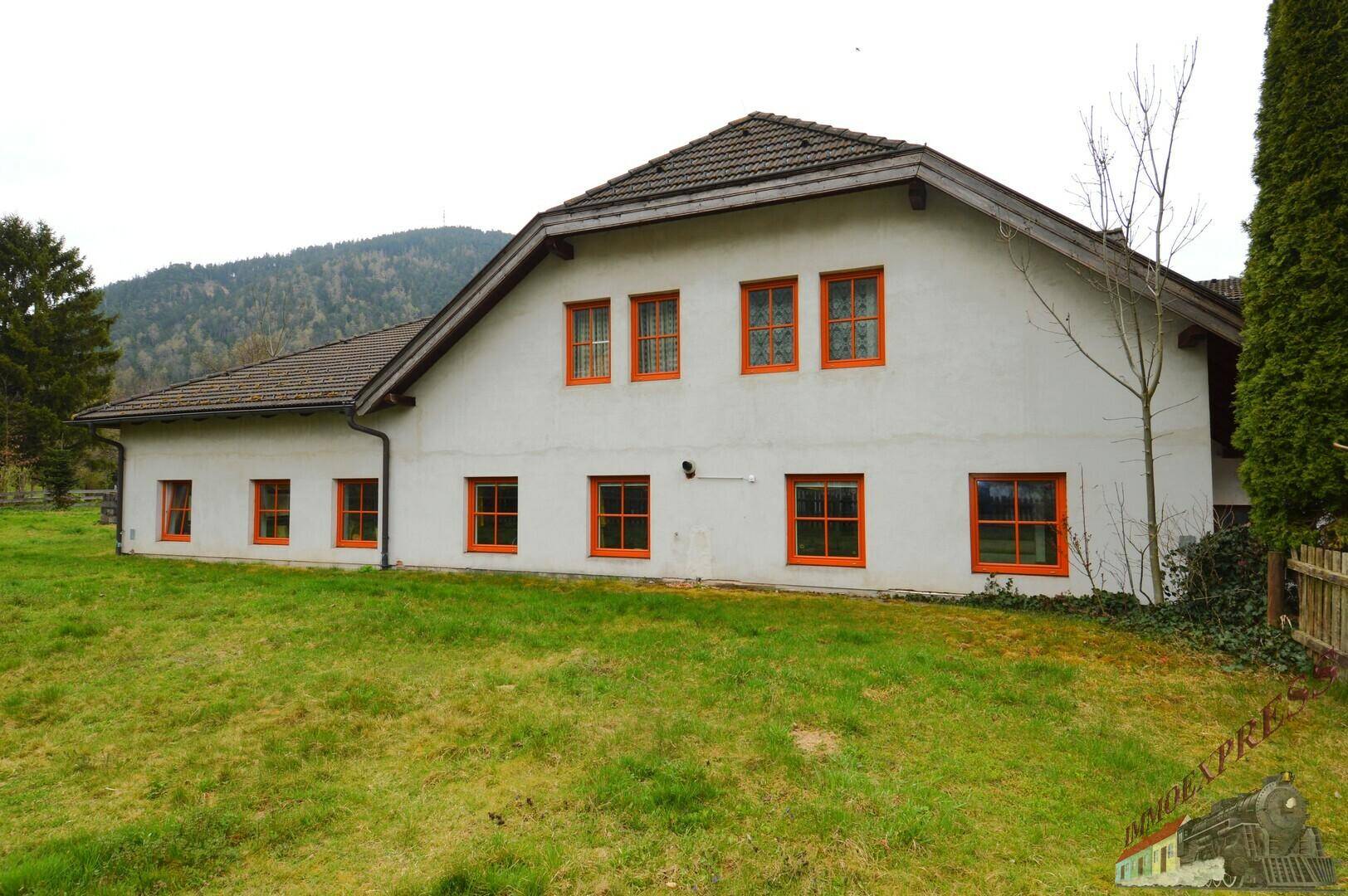 Weißes Haus mit orangefarbenen Fensterrahmen auf einem grünen Hügel mit Bergblick.