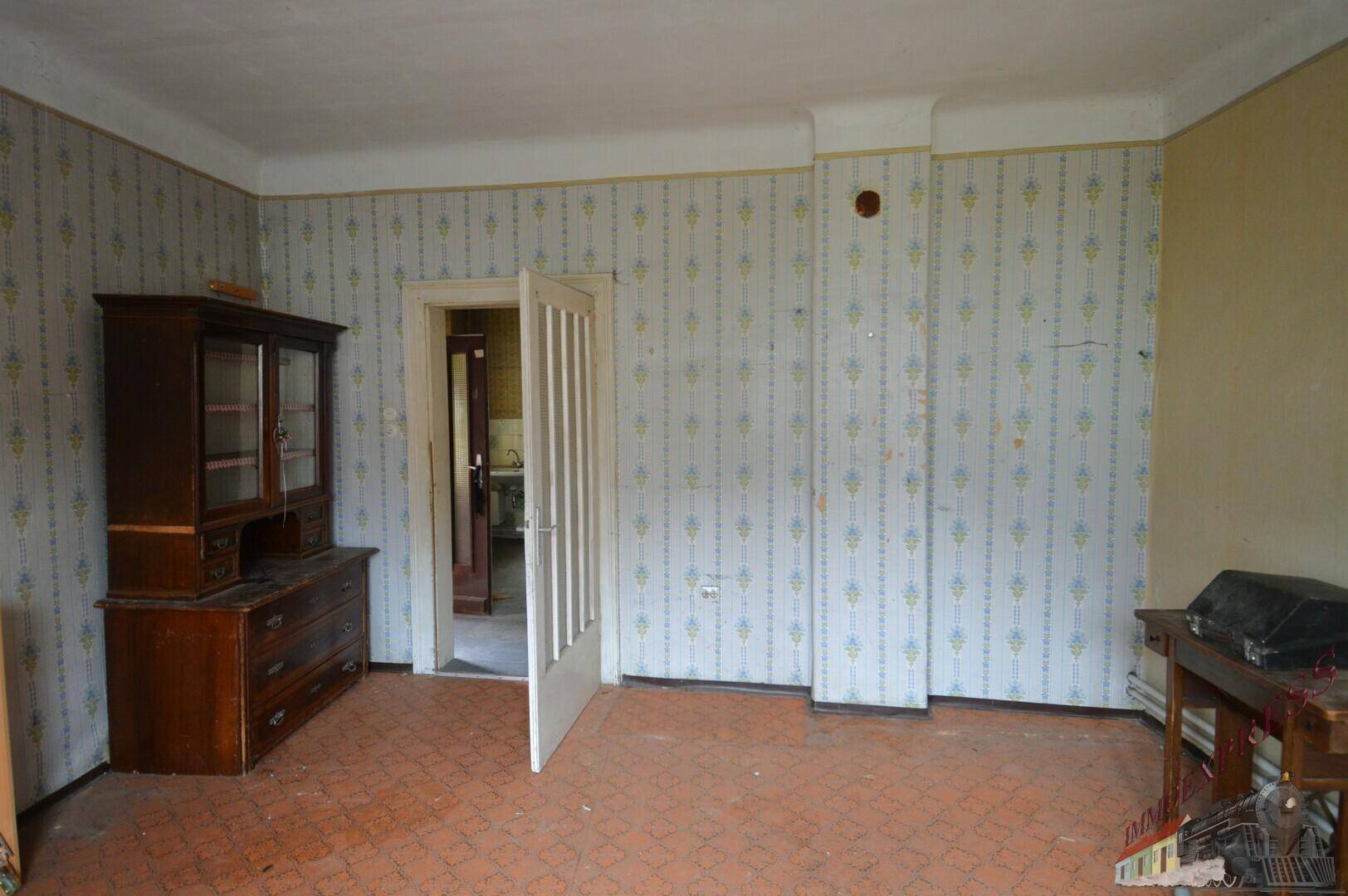 Wohnraum mit gemusterter Tapete und einem antiken Schrank, der auf eine Renovierung wartet.