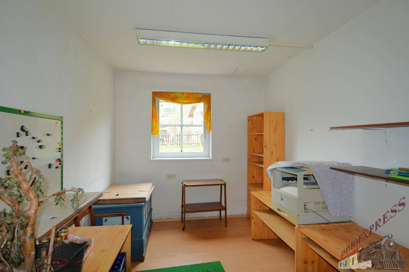 Einfaches Arbeitszimmer mit Fensterblick ins Grüne, Holzregalen und funktionaler Einrichtung.
