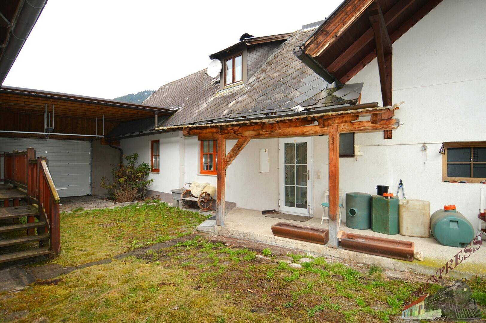 Ländliches Gebäude mit überdachtem Eingangsbereich, Garage und einem ungepflegten Garten.