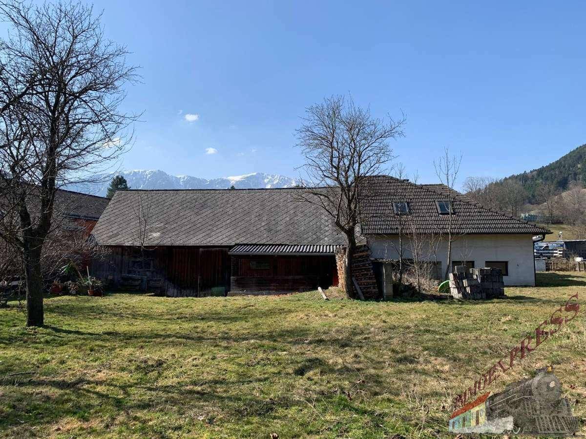 Ländliches Anwesen mit Wohnhaus und Nebengebäude, umgeben von einer weiten Wiese und Bergblick im Hintergrund.