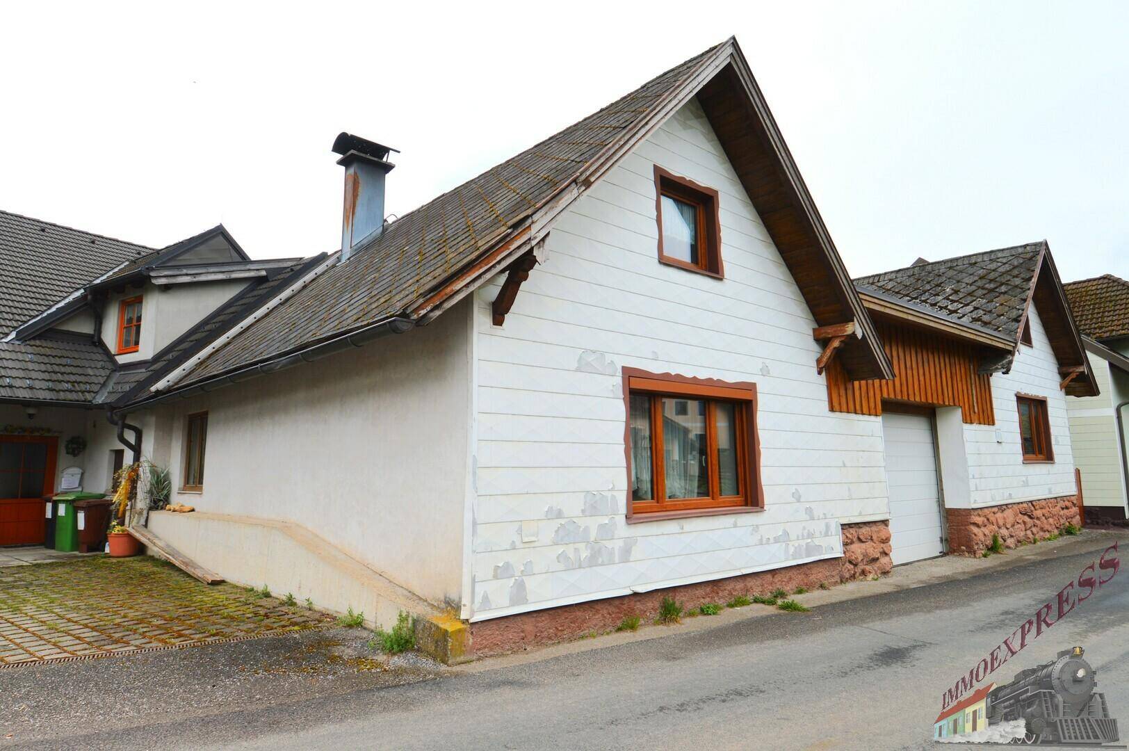 Seitenansicht eines ländlichen Hauses mit weißer Fassade, Holzverkleidung und Zufahrt von der Straße.