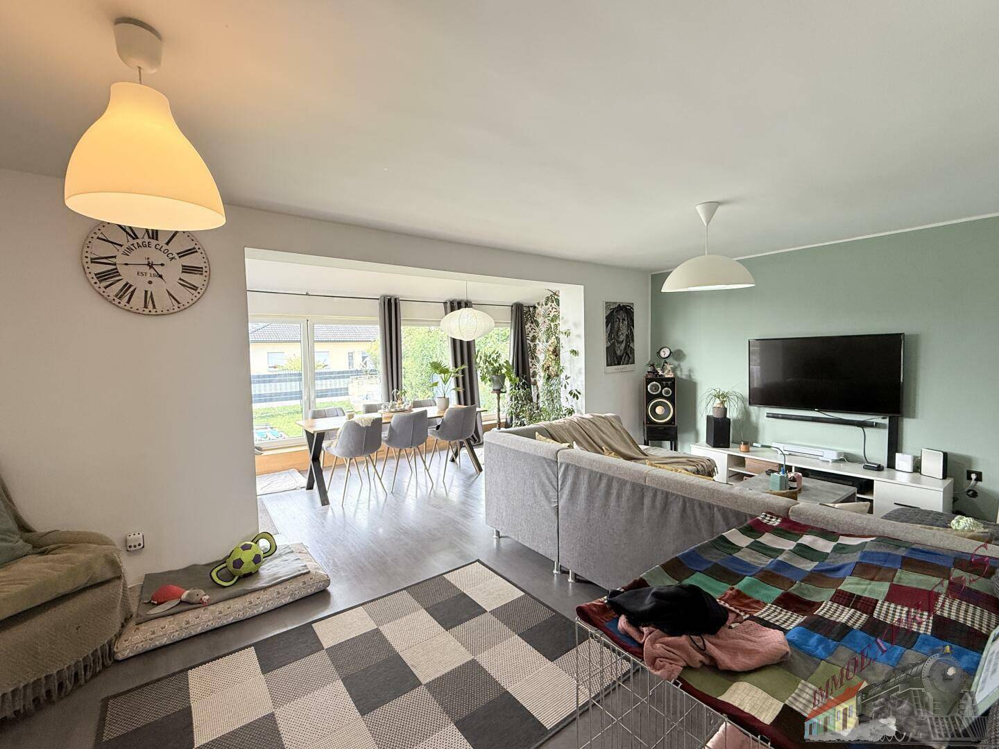 Offener Wohn- und Essbereich mit modernem Sofa, Fernseher und großem Esstisch am Fenster.