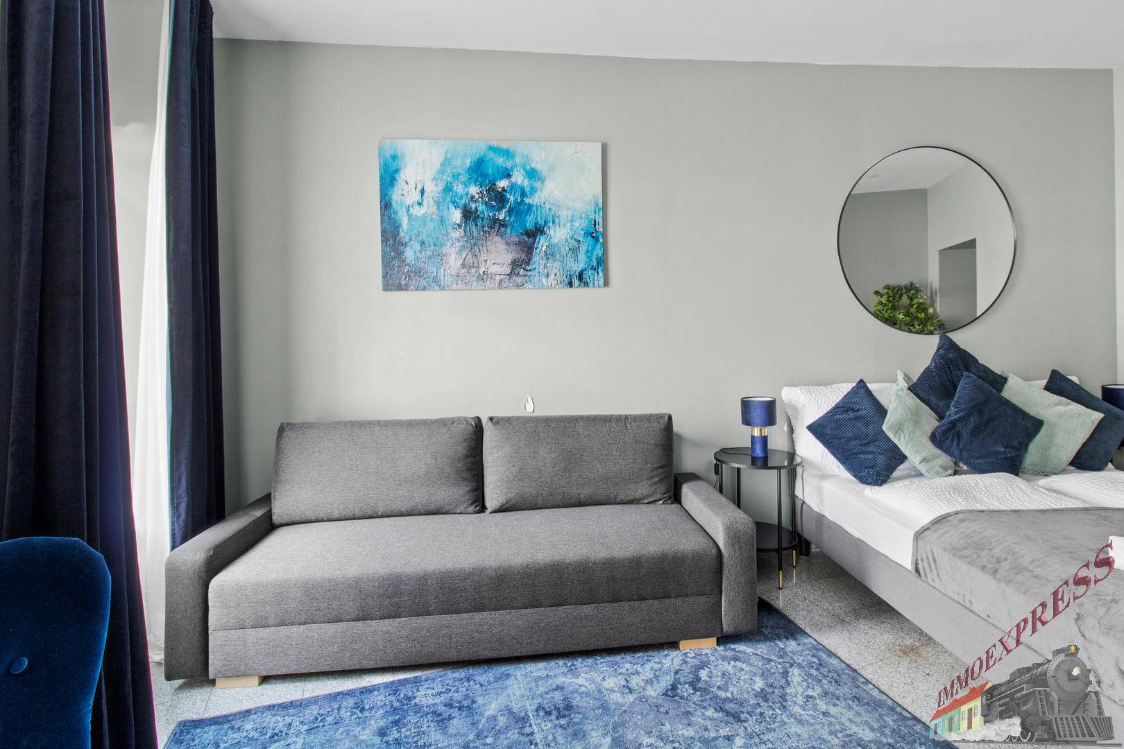 Schlafzimmer mit einem bequemen Schlafsofa, blauen Teppichen und einem runden Wandspiegel.