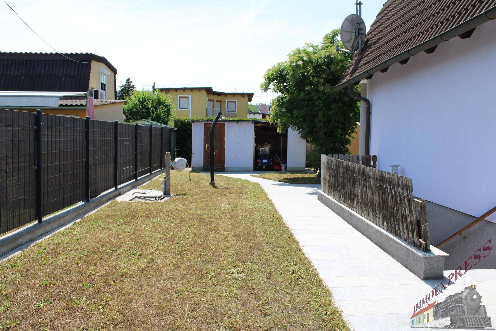 Hinterhof mit gepflastertem Weg, Gartenhaus und einem hohen Sichtschutzzaun.