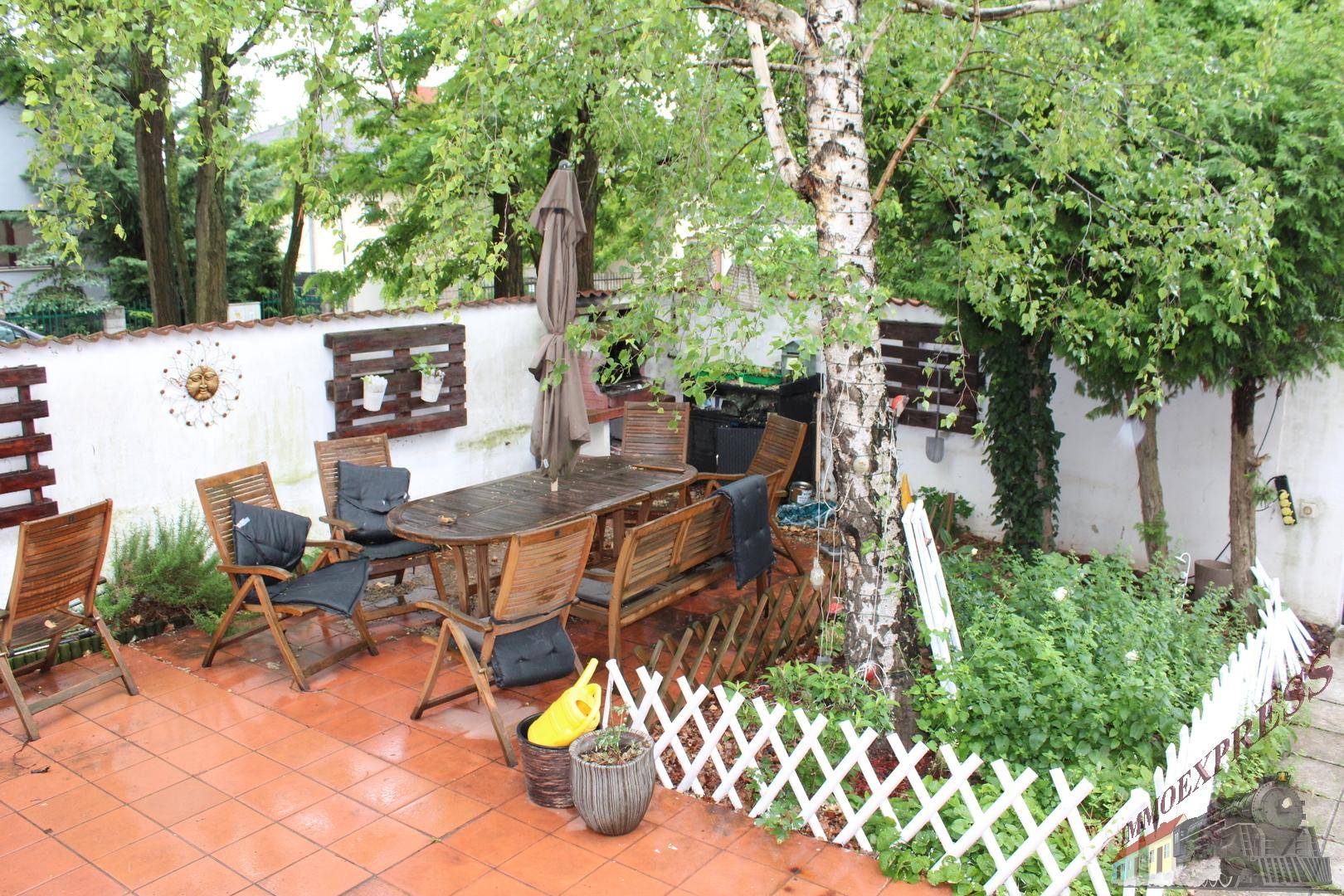 Gemütliche Terrasse mit Holztisch und Stühlen, umgeben von Pflanzen und einem kleinen Zaun.