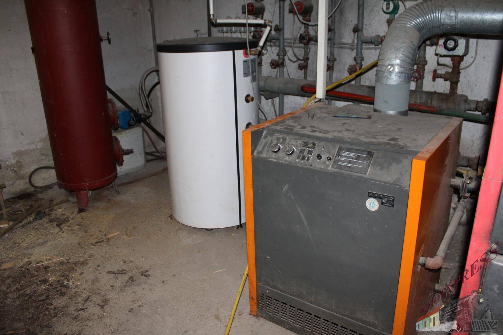 Heizungsanlage im Keller mit Kessel und Warmwasserspeicher, umgeben von Rohren.
