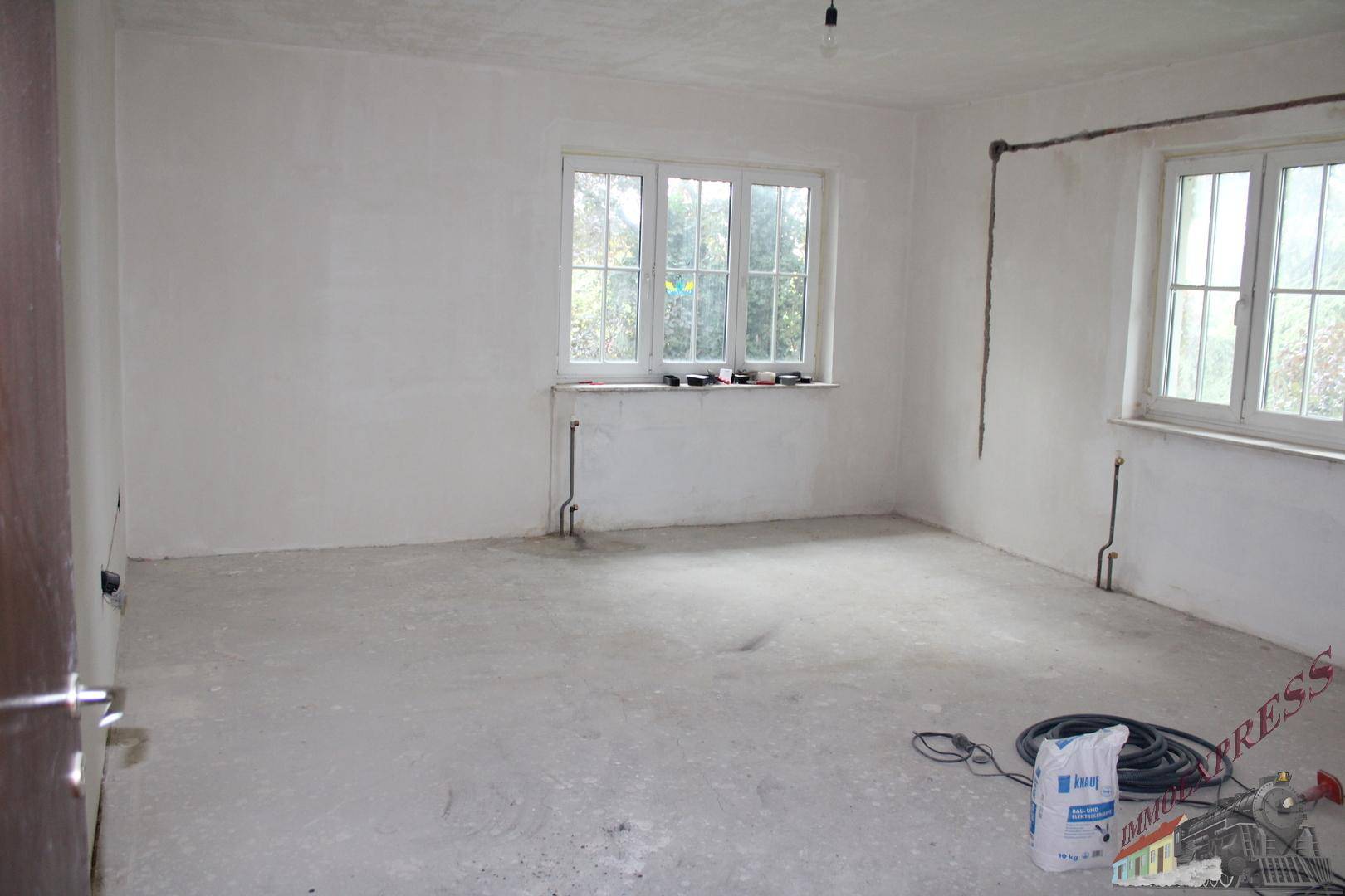 Leerer Raum im Rohbauzustand mit Fenstern und vorbereiteten Installationen.