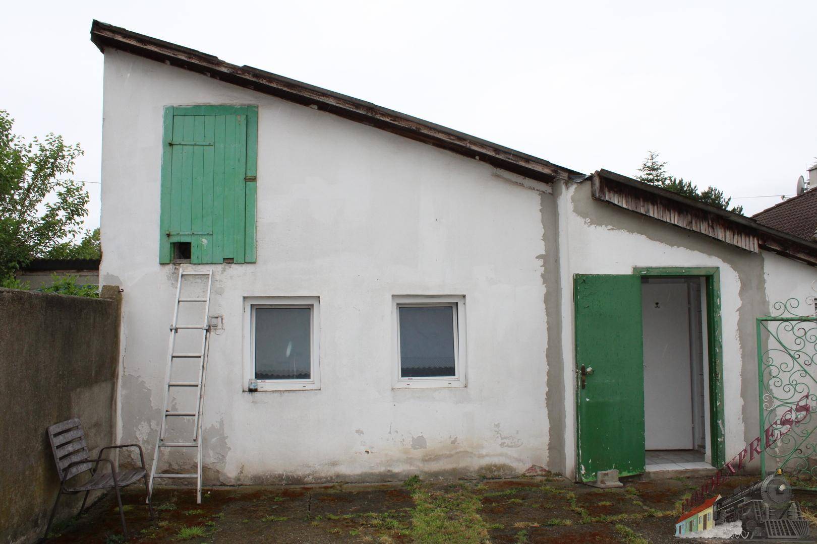 Außenansicht eines einfachen Nebengebäudes mit weißer Fassade, grünen Türen und einem Pultdach.