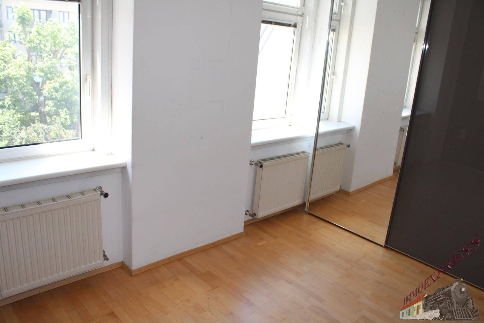 Helles Zimmer mit Holzboden, Spiegelschrank und Blick ins Grüne durch die Fenster.