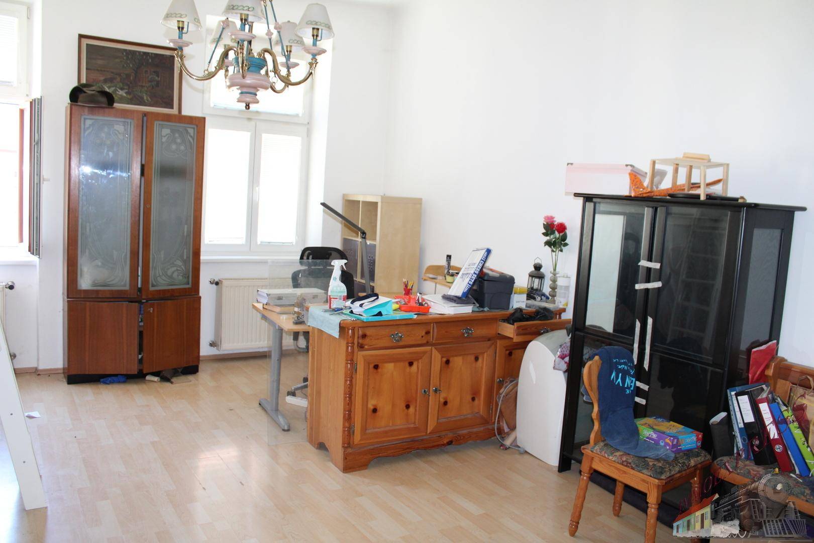 Geräumiges Zimmer mit Holzboden, Schreibtisch und verschiedenen Möbelstücken unter einem Kronleuchter.