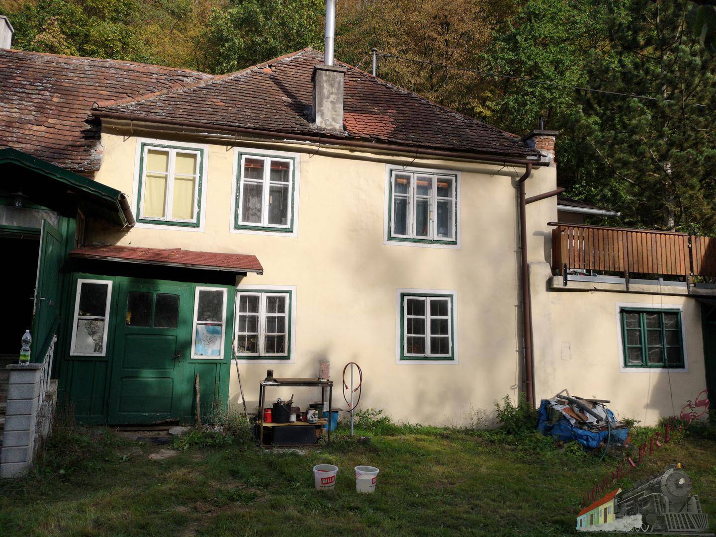 Rückseite eines ländlichen Hauses mit kleinem Holzbalkon und grüner Umgebung.